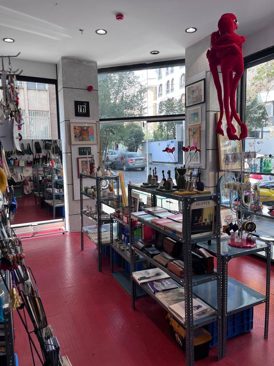 Inside the store by day: red floor, steel shelves of books, leather cases, candles and jewelry, framed art on the pillars