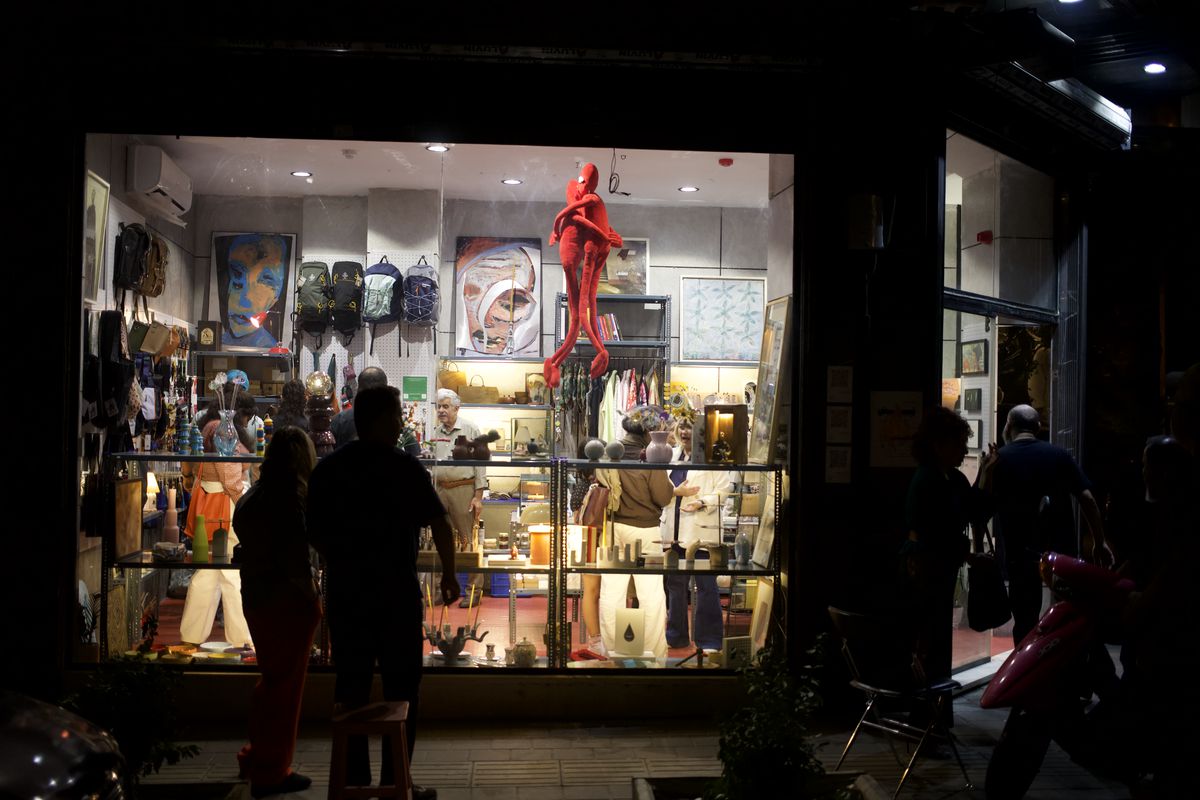 The TASH collective shop window lit up at night at the corner of Abdoh and Sarbedaran, with paintings, backpacks, ceramics and a large red plush figure hanging inside